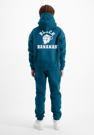 Persoon draagt een teal hoodie en bijpassende broek met witte sneakers, achterkant van de hoodie toont de tekst "Black Bananas" en het apenlogo.