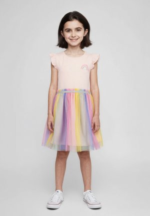 Robe courte rose à manches courtes avec des épaules à volants, ornée d'un patch arc-en-ciel et d'une jupe en tulle multicolore. Associée à des baskets blanches.