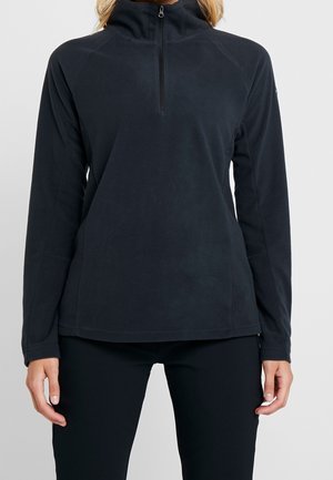 Vrouw die een zwarte fleece-pullover met lange mouwen, een korte voorrits en een hoge kraag draagt, staand tegen een effen achtergrond.
