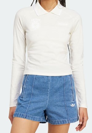 Femme portant une chemise blanche à manches longues avec col et un short en jean bleu taille haute avec le logo Adidas sur la jambe gauche.