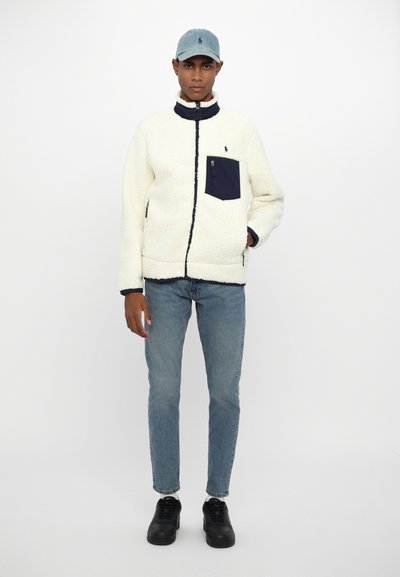 Fleece jacket in cream with a navy zippered pocket and collar, paired with light denim jeans and black sneakers. Cap is light blue.