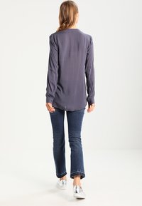 Blouse grise à manches longues avec une texture lisse et des poignets boutonnés, associée à un jean bleu foncé évasé et des baskets argentées.