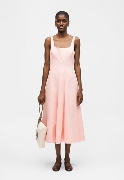 Vrouw die een lichtroze mouwloze midi-jurk draagt, een witte handtas met bruine band vasthoudt, bruine platte schoenen draagt, staand tegen een witte achtergrond.