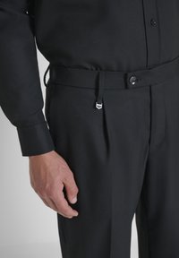 Muškarac nosi crne hlače i usklađenu crnu košulju s dugmetom na struku i detaljem od srebrnog remena.