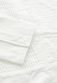 Valge särk, millel on tekstuuriga kangas, vertikaalsed voltid, nööpidega mansetid ja selged plastiknööbid kinnitamiseks.