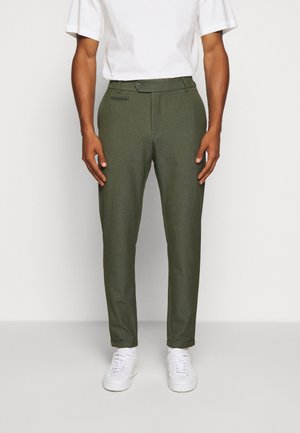 Homme portant un pantalon slim vert olive, un t-shirt blanc col rond et des baskets blanches, debout devant un fond clair uni.
