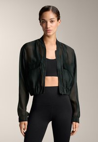 Musta lyhyt bomber-takki, jossa on läpikuultavaa kangasta, pitkät hihat, joustavat hihansuut ja vetoketjukiinnitys edessä. Yhdistetty mustiin istuviin leggingsseihin.