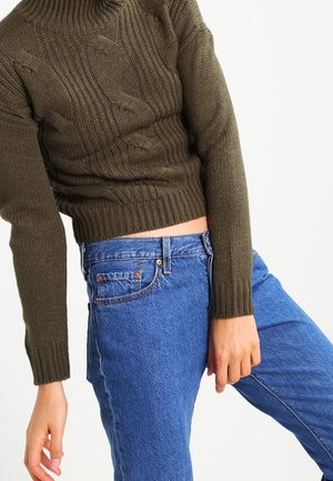 Osoba ubrana w brązowy dziergany sweter i niebieskie, wysokowaistowe dżinsy, lekko pochylona na bok, z jedną ręką swobodnie opuszczoną.