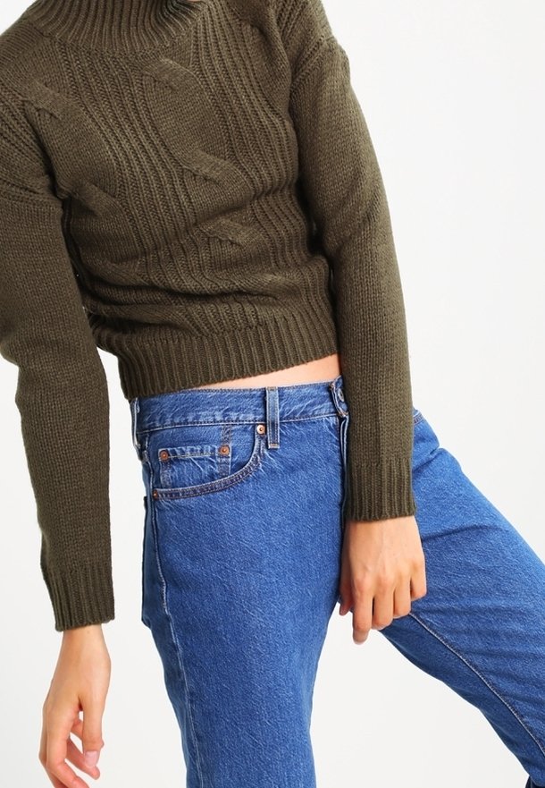 Personne portant un pull en tricot marron et un jean bleu taille haute, légèrement penchée sur le côté avec une main détendue vers le bas.