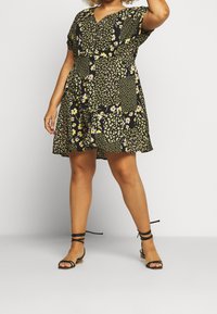 Schwarzes Blumenkleid mit kurzen Ärmeln, das mit Patchwork-Mustern in Gelb und Grün versehen ist. Kombiniert mit schwarzen Sandalen mit Riemen und einem niedrigen Blockabsatz.