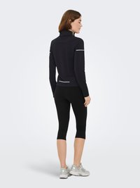 Zwart atletisch topje met een hoge kraag, voorzien van witte reflecterende accenten, gecombineerd met zwarte capri-legging en lichtgrijze sneakers.