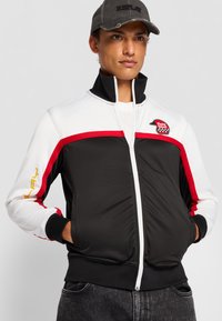 Black and white zip-up jacket with red and yellow accents, high collar, and front pockets. Features logo patch on the chest.