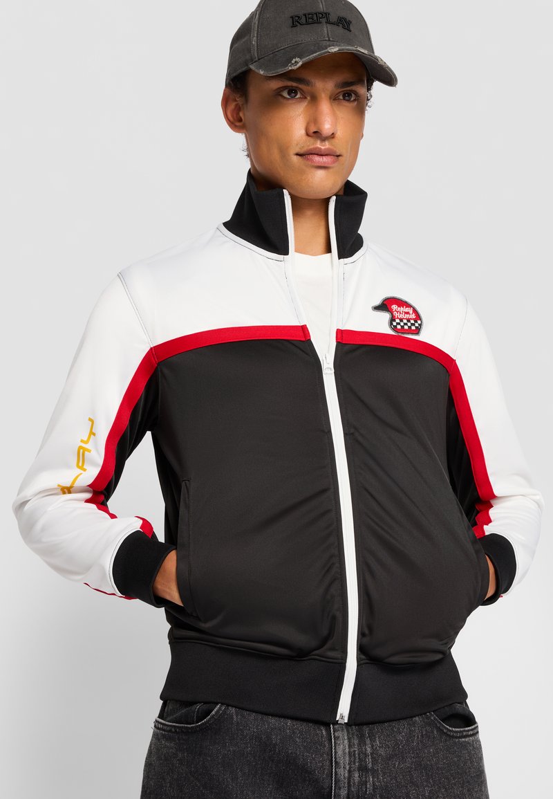 Schwarz-weiße Zip-Jacke mit roten und gelben Akzenten, hohem Kragen und Fronttaschen. Verfügt über ein Logo-Patch auf der Brust.