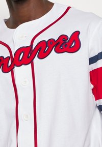 Wit honkbalshirt met rode en navy strepen op de mouwen en "Braves" in rode letters met navy omlijning over de borst.