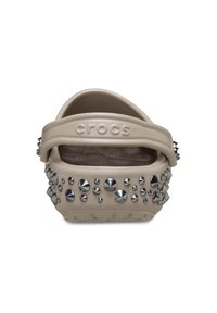 Beige Crocs mit einer strukturierten Fußbett, die metallische silberne Spitzen und Nieten an den Seiten aufweisen. Verstellbarer Riemen hinten für einen sicheren Sitz.