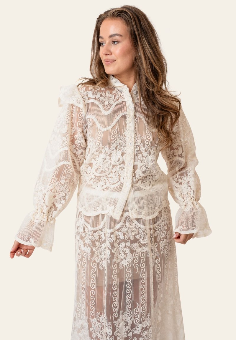 Femme aux longs cheveux bruns porte un chemisier en dentelle blanche transparente et une jupe à motifs floraux et rayés, debout devant un fond clair uni.
