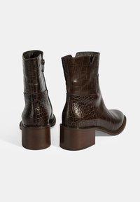 Braune Lederstiefeletten mit Krokodilprägung, runder Zehenform und robustem Absatz. Mit einem Reißverschluss an der Seite für ein einfaches An- und Ausziehen.