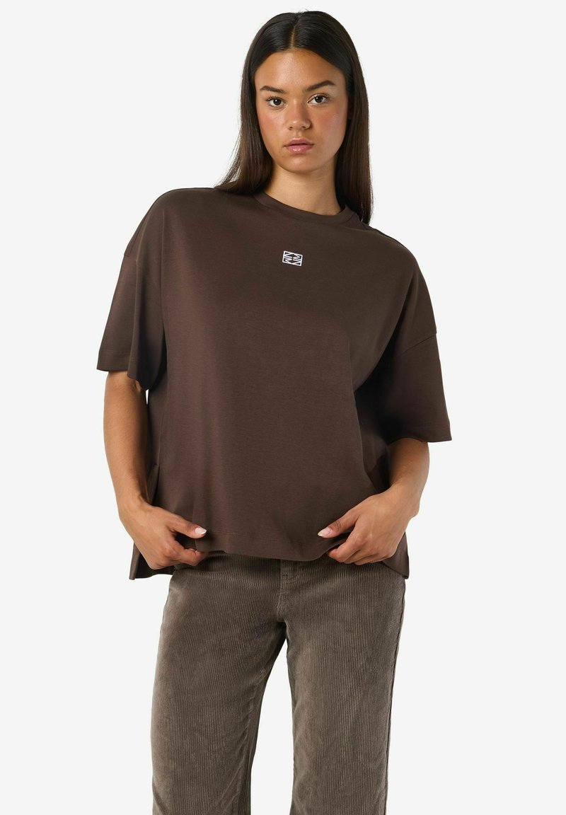 Brązowy, oversize t-shirt wykonany z miękkiej bawełny, z krótkimi rękawami i naszywką z logo na piersi. W zestawie z jasnobrązowymi spodniami z sztruksu.