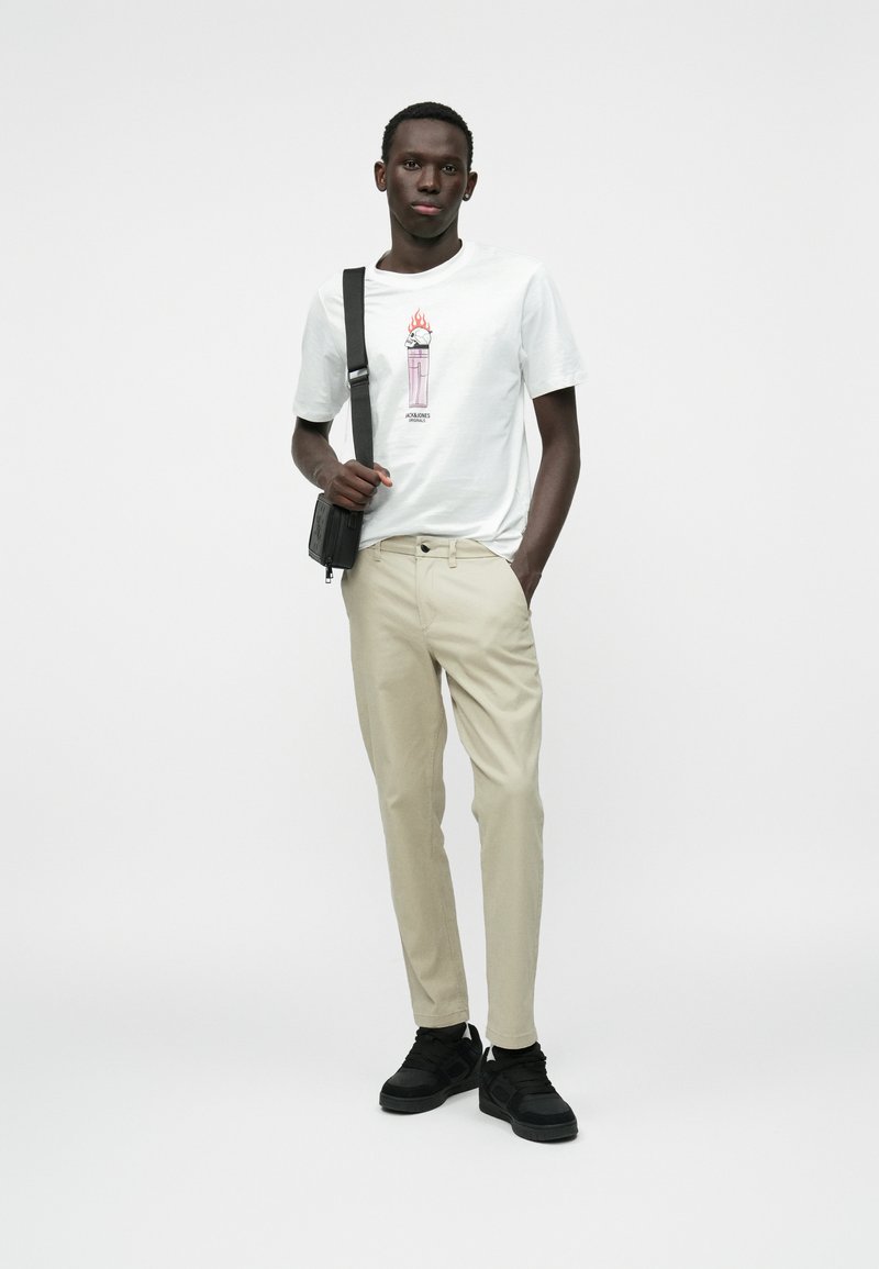 Biely tričko s grafickým potlačou, béžové nohavice a čierne tenisky. Doplnkom je čierna crossbody taška. Jednoduchý dizajn a ležérny strih.