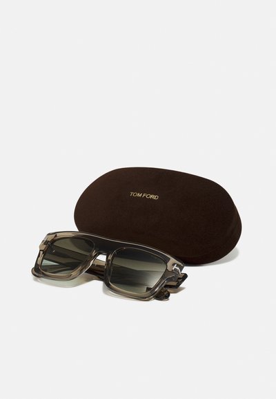 Ochelari de soare Tom Ford cu o ramă gri transparent și lentile închise, însoțiți de un toc protector din suede maro. Logo-ul auriu pe toc.