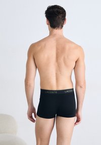 Modelo masculino de costas para a câmara, vestindo boxers pretos da Lacoste, contra um fundo claro e liso.