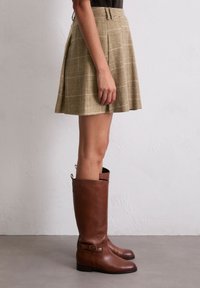 Une jupe patineuse à carreaux beige avec des plis présente une texture lisse. Associée à de hautes bottes en cuir marron avec un accent à boucle.