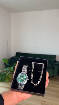 Main tenant une boîte noire contenant une montre en métal argenté avec un cadran vert et un bracelet chaîne assorti en argent.