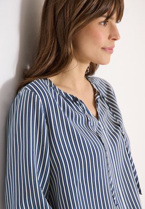 Femme vêtue d'un chemisier rayé bleu et blanc à rayures verticales, tournée vers la droite sur un fond blanc, montrant un profil de côté et des cheveux bruns mi-longs.