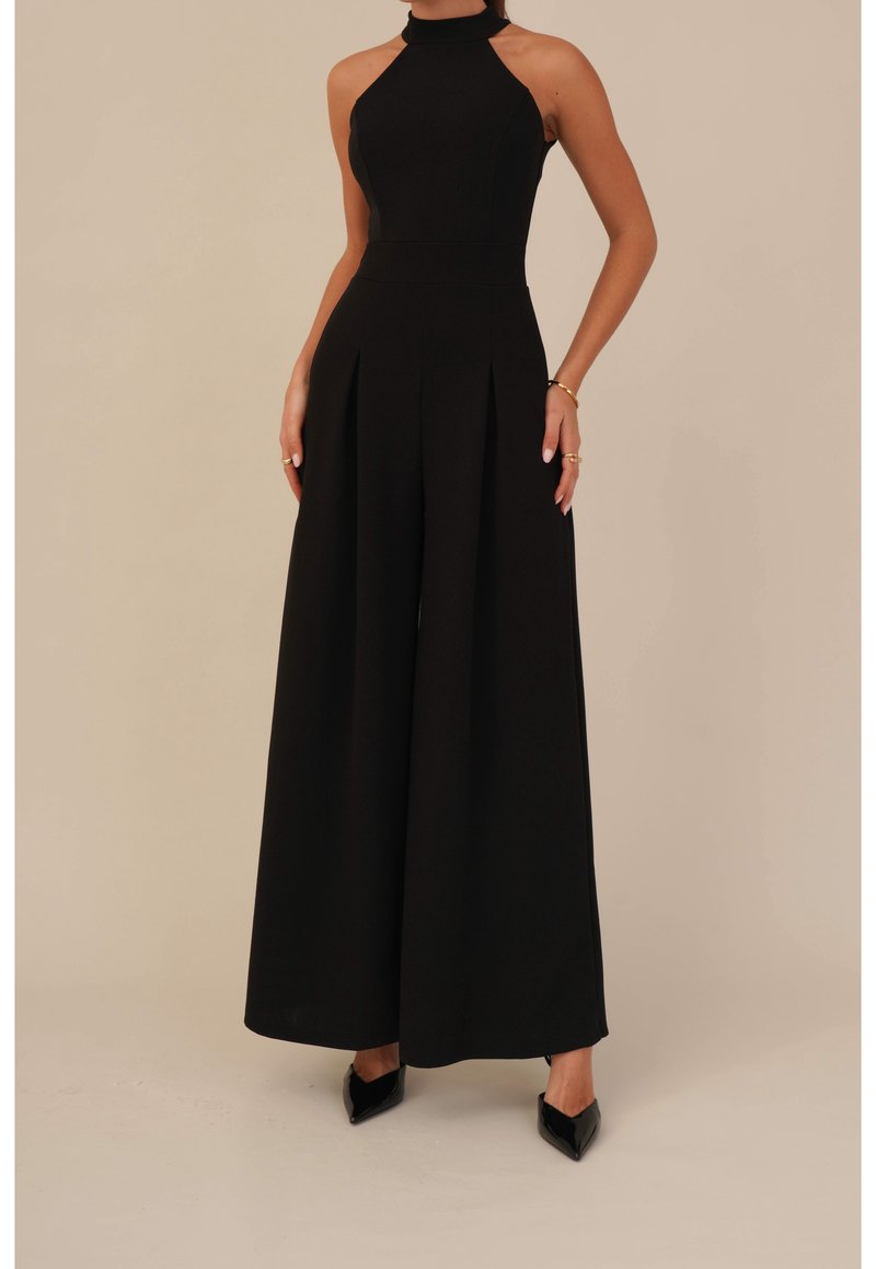 Femme portant une combinaison noire sans manches col halter avec jambes larges et talons noirs à bouts pointus, debout, les mains le long du corps.