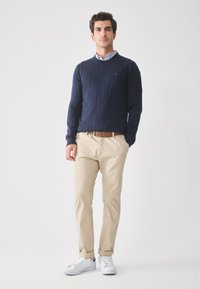 Homme debout portant un pull côtelé bleu marine, une chemise à col bleu clair, un pantalon beige avec une ceinture marron et des baskets blanches sur fond blanc.