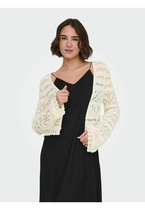 Kobieta w czarnej sukience bez rękawów, nosząca biały ażurowy sweterek na szydełku, z krótkimi falowanymi włosami i złotymi kolczykami-kołami.