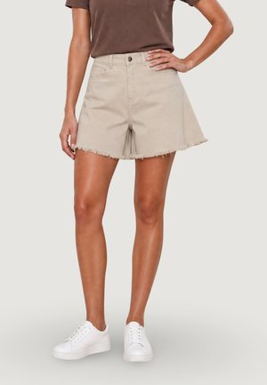 Donna che indossa pantaloncini beige con orlo sfrangiato, camicia marrone e scarpe da ginnastica bianche, in piedi con una mano sul fianco davanti a uno sfondo neutro.