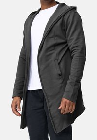 Chaqueta capucha gris oscuro con mangas largas, corte asimétrico y bolsillos frontales. Confeccionada en tejido texturizado, con bordes sin rematar.