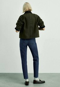 Veste courte vert olive avec un col noir, associée à un jean bleu et des mocassins marron. La tenue se caractérise par des manches retroussées et des chaussettes blanches.