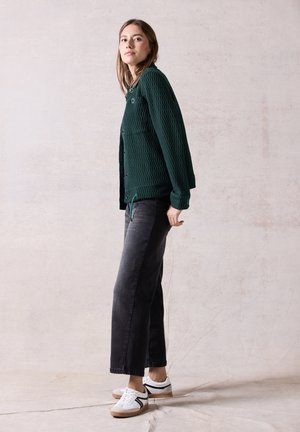 Giacca a maglia verde con texture e tasche anteriori, abbinata a jeans neri a gamba larga e sneakers bianche con dettagli neri.