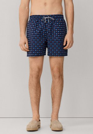 Marineblaue Badeshorts mit kleinen roten und hellblauen Mustern, Kordelzug in der Taille, getragen mit beigen Slip-on-Espadrilles.