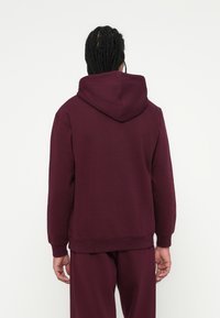 Zsinóros hajú személy burgundi színű kapucnis pulóverben és hozzá illő nadrágban áll háttal egy sima fehér háttér előtt.