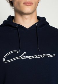 Sudadera con capucha de color azul marino, hecha de un tejido suave, que presenta el texto cursivo blanco "Closure" en el frente, con ojales plateados para los cordones.