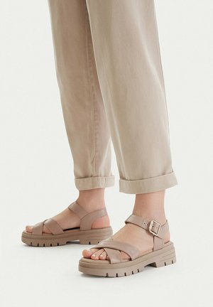 Piedi che indossano sandali con suola spessa beige e cinturini alla caviglia, con pantaloni beige chiaro risvoltati, in piedi su una superficie bianca.