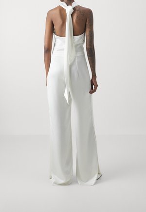 Persoon met tatoeages die een wit haltertop zonder rug draagt, vastgebonden in de nek, en bijpassende wijde broekspijpen, staande tegen een effen achtergrond.