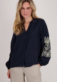 Donkerblauw, knoopjesoverhemd met een V-hals, uitgerust met opgeblazen mouwen en gedetailleerde lichtgekleurde borduursels op de linker mouw. Gladde stofstructuur.