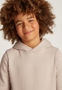 Bambino sorridente con capelli ricci che indossa un hoodie Calvin Klein Jeans beige chiaro su uno sfondo semplice e chiaro.