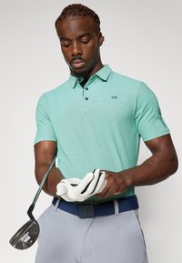 Grünes, strukturiertes Poloshirt mit Kragen und drei Knöpfen, kombiniert mit grauen Hosen und einem blauen Gürtel. Ein Golfschläger und ein Handschuh werden in einer Hand gehalten.