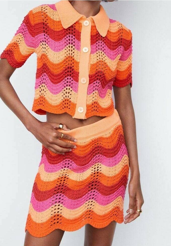 Femme portant un ensemble en tricot coloré à deux pièces avec des rayures ondulées en orange, rouge, rose et p êche, comprenant un haut court à boutons et une jupe.