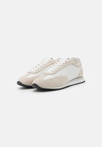 Beige suedesko og hvite skinnsko med en glatt tekstur, flate lisser og en svart gummisåle. Rund tå og minimalistisk design.