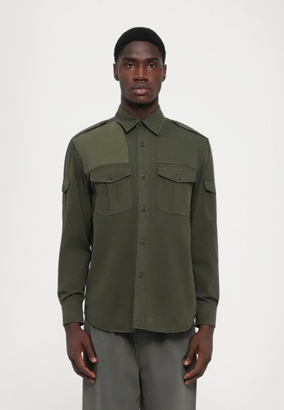 Chemise olive vert à manches longues avec deux poches poitrine, boutonnière sur le devant et épaulettes. Tissu lisse avec un design structuré.