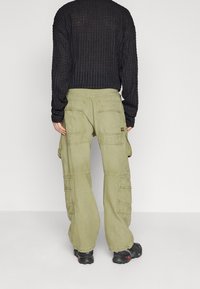 Person wearing loose-fitting olive green cargo pants with multiple back and side pockets, black textured long-sleeve top, and black shoes.