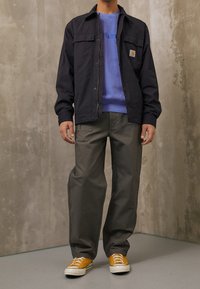 Muž v černé jacket, modré mikině, šedých kalhotách a žlutých teniskách stojí proti texturované šedé zdi.
