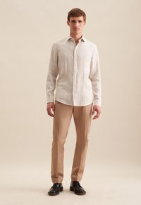 Camicia a maniche lunghe a righe in beige chiaro e bianco, con bottone, abbinata a pantaloni beige e scarpe nere, in posizione eretta.