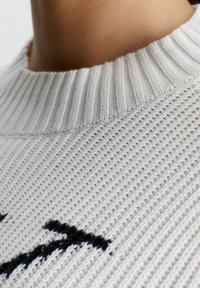 Maglione grigio chiaro a maglia con texture, caratterizzato da un collo a coste e un design nero. Il materiale sembra morbido e traspirante con un motivo tessuto.